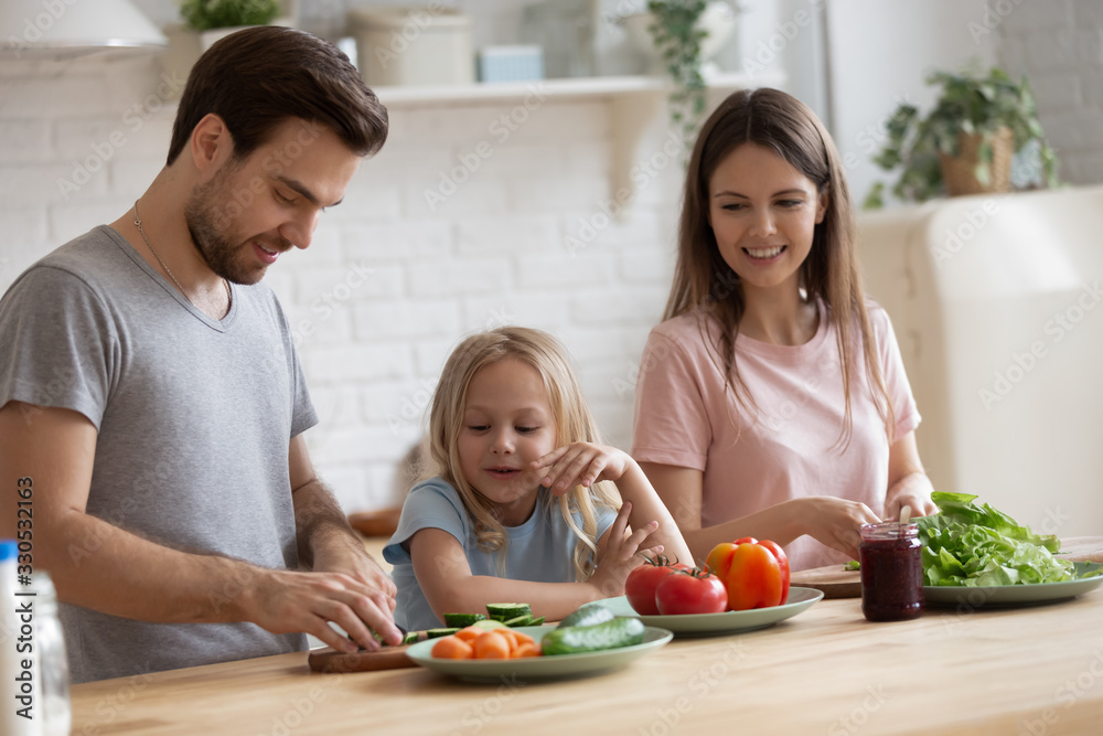 Smiling young parents have fun chopping vegetables preparing salad or lunch with cute little daughter, happy mom and dad cooking together with small preschooler girl child, healthy lifestyle concept