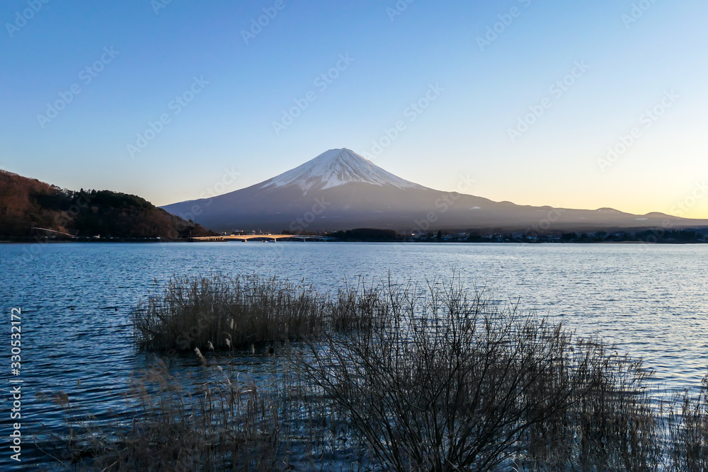 A close up view on Mt Fuji from the side of Kawaguchiko Lake, Japan. Soft colors of sunset - golden hour. Top of the volcano covered with a snow layer. Serenity and calmness. Calm lake's surface