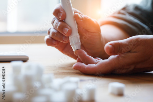 senior woman hands using lancet on finger at home to check blood sugar level, glucometer  and sugar cubes on wooden table close up, diabetes concept,  elderly health care, sunny morning photo