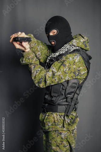Obraz na plátně A man in camouflage uniform, body armor and a balaclava, holds his weapon ready and takes aim with a pistol, standing against a gray background