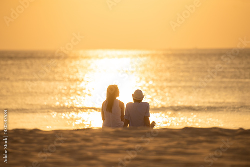 couple sit on sand looking sunset in sea together.