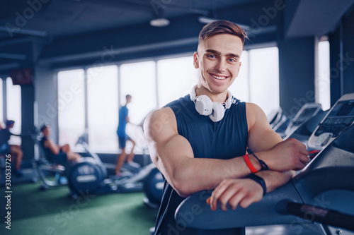 Smiling positive confident male personal instructor with arms crossed arms near treadmill at gym in fitness gym.