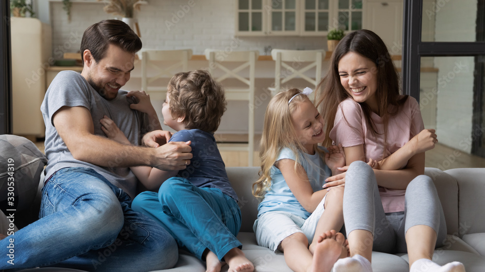 Playful young parents have fun play with happy little kids tickle laugh sitting on couch in living room, overjoyed family enjoy funny childish activity or game with small children on weekend at home Playful young parents have fun play with happy little kids tickle laugh sitting on couch in living room, overjoyed family enjoy funny childish activity or game with small children on weekend at home