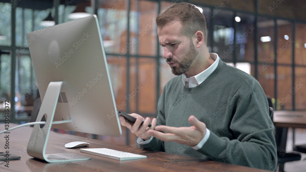 Angry Man get Call on Smartphone while working on Computer Angry Man get Call on Smartphone while working on Computer