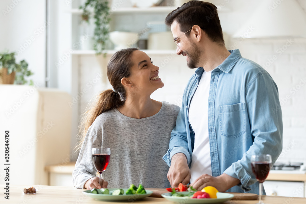 Happy young couple cooking dinner in kitchen at home together, smiling husband cutting fresh vegetables, preparing salad, beautiful wife holding wine glass, enjoying romantic moment, having fun