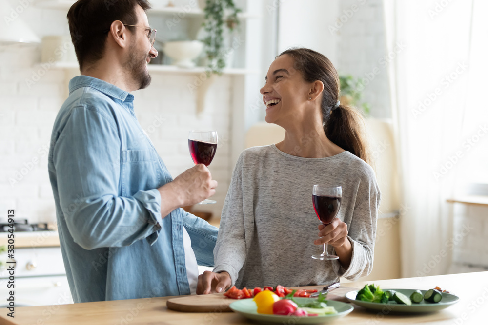 Young loving couple having fun in kitchen, drinking red wine, holding glasses, laughing beautiful wife and husband chatting, preparing salad, cooking dinner, enjoying free time together - obrazy, fototapety, plakaty 