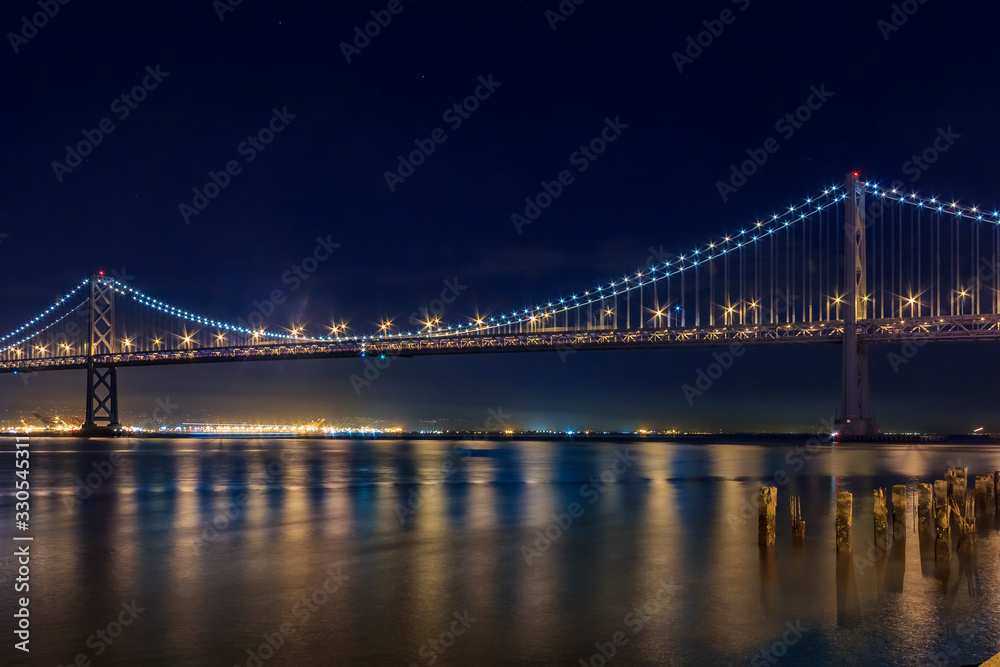 Fototapeta premium San Francisco Bay Bridge w nocy oświetlony żółtymi i niebieskimi światłami odbijającymi wodę w zatoce, długi czas naświetlania