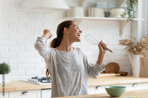 Excited funny girl singing into whisk, having fun in modern kitchen at home, happy girl holding beater as microphone, dancing, listening to music, having fun with kitchenware, preparing breakfast photo
