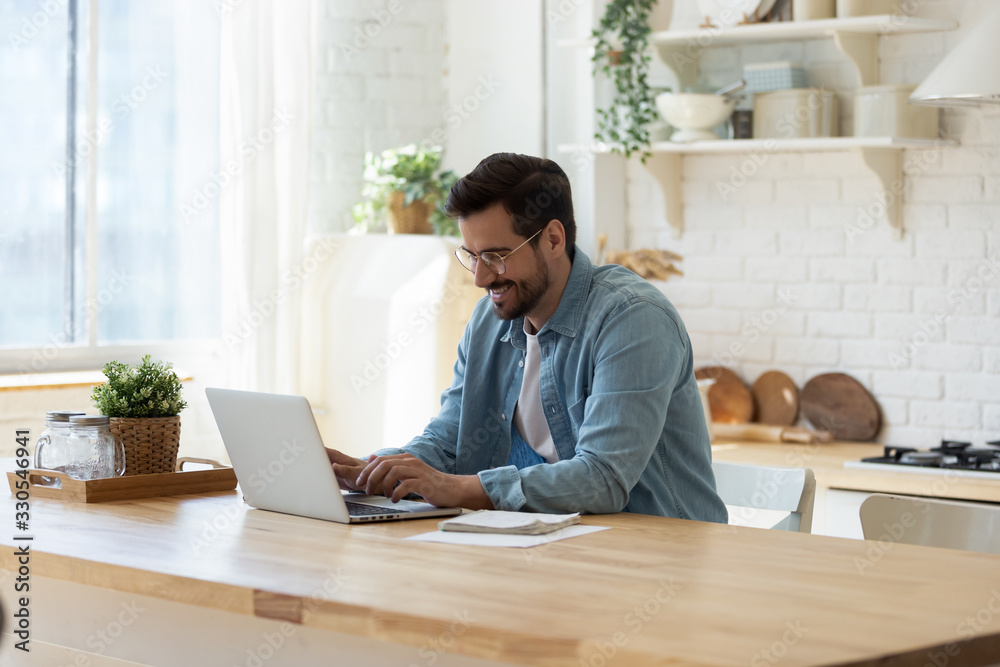 Smiling young man working on laptop in modern kitchen, checking email in morning, writing message in social network, happy young male using internet banking service, searching information