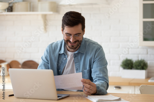 Excited young man wearing glasses reading good news in letter, sitting at table with laptop, happy satisfied male holding document, paper notification, job promotion, loan approval, great exam result