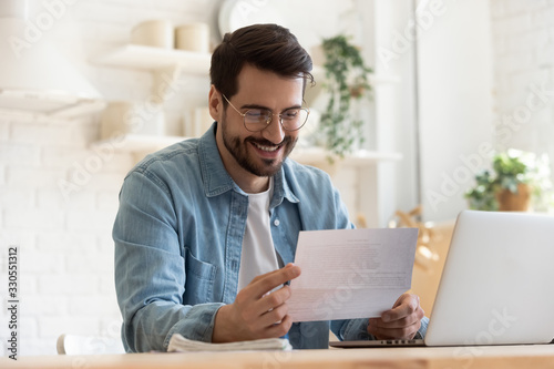 Smiling young man reading good news in paper notification, sitting at table with laptop, happy satisfied male wearing glasses holding document, job promotion, loan approval, great exam result photo