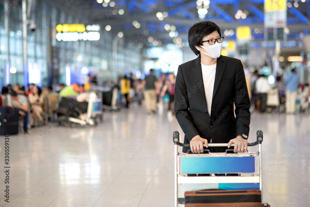Fototapeta premium Asian businessman wearing suit and face mask walking with airport trolley and suitcase luggage in airport terminal. Coronavirus (COVID-19) outbreak prevention. Health awareness for pandemic protection