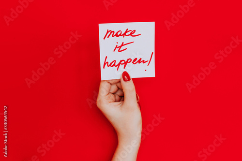 Note in a female hand with manicure, red nails. "Make it happen!" sign. Background red