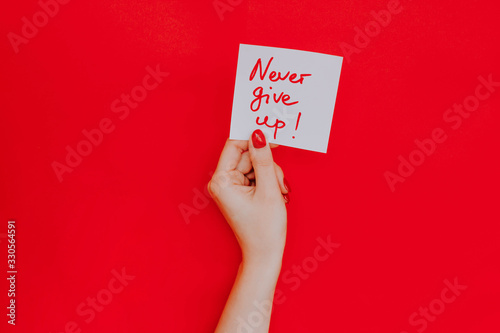 Note in a female hand with manicure, red nails. "Never give up!" sign. Background red