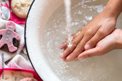 Child’s hands under white bowl with water upon water stream, colorful soaps on a white material, cleanliness and hygiene concept 