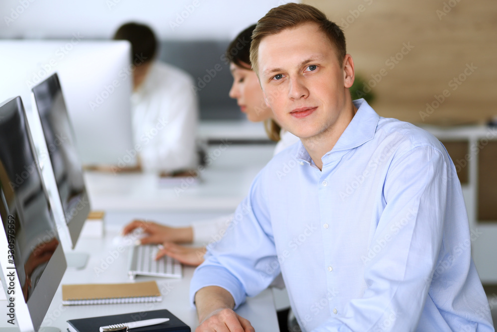 Fototapeta premium Cheerful smiling businessman headshot at work in modern office. Unknown casual dressed entrepreneur using pc computer while sitting with diverse colleagues at the background. Multi ethnic working