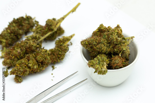 White porcelain dish full of dried cannabis buds. Rest of weed laying on white table. Marijuana for medicinal purpose. Medicine, drugs and health care concept...