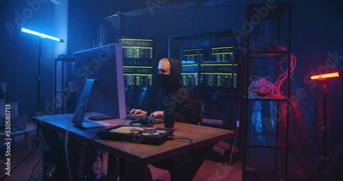 Caucasian anonymous woman with short blond hair and mask on face working in cyber security center and stealing data. Female hacker typing on keyboard at big computer in dark monitoring room. photo