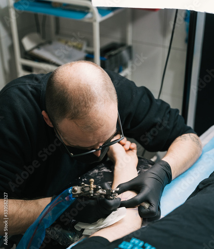 A closeup shot of a tattoo artist with eyeglasses creating a tattoo on a man's arm