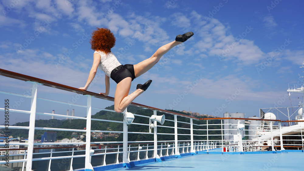 Fototapeta premium Slim beautiful girl dancer on observation deck cruise ship jumping dance elements. beautiful lines of red-haired curly-haired European girl on blue podium, background of an Asian city. modern dance