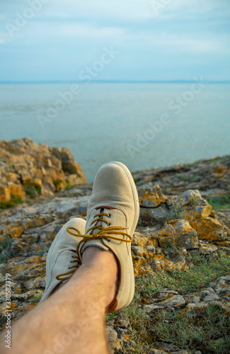Feet of male tourist resting on the rocks at the beach at sunset, looking at the sea.