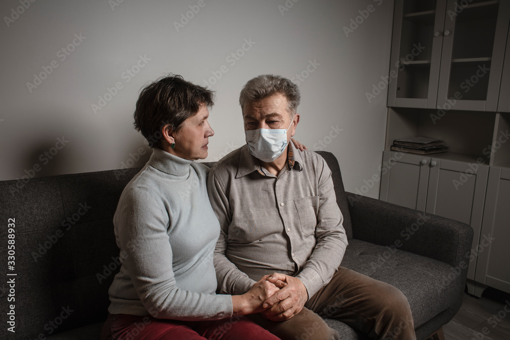 A couple sitting on a sofa wearing a protective mask. An infectious agent protection gear  including a mask. An sick older man and woman wearing protective masks to protect against virus. Coronavirus.