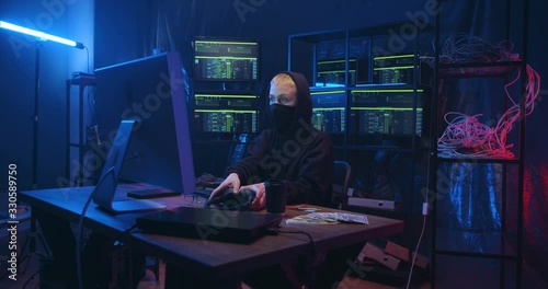 Serious Caucasian woman in headphones and mask sitting in front of big screen of computer while creating virus program. Female anonymous cyber criminal with closed face stealing data at online server. photo