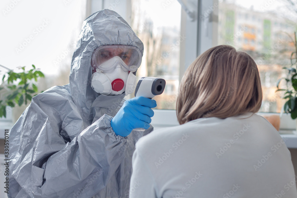 Sprawdzanie temperatury za pomocą termometru laserowego przez osobę w kombinezonie ochronnym, masce ochronnej oraz okularach. Termometr trzymany w dłoniach ubranych w rękawiczki jednorazowe.