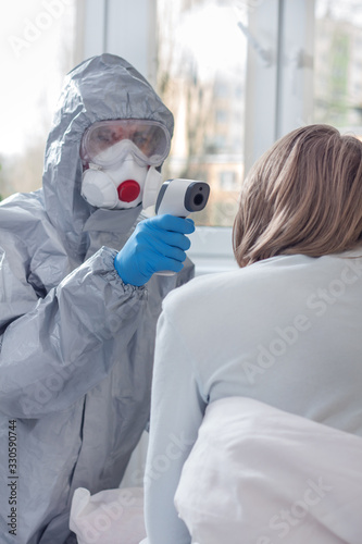 Sprawdzanie temperatury za pomocą termometru laserowego przez osobę w kombinezonie ochronnym, masce ochronnej oraz okularach. Termometr trzymany w dłoniach ubranych w rękawiczki jednorazowe.