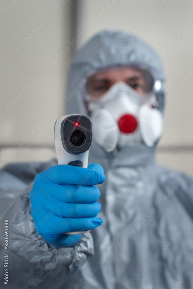 Na pierwszym planie dłoń ubrana w rękawiczkę jednorazową trzyma termometr laserowy i mierzy temperaturę. W tle postać ubrana w kombinezon ochronny, maskę na twarz i okulary ochronne.
