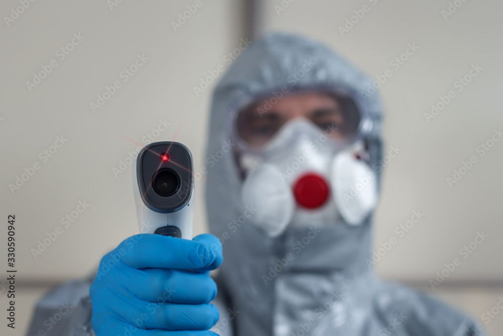 Na pierwszym planie dłoń ubrana w rękawiczkę jednorazową trzyma termometr laserowy i mierzy temperaturę. W tle postać ubrana w kombinezon ochronny, maskę na twarz i okulary ochronne.