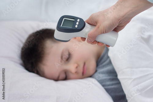 Na pierwszym planie dłoń trzyma termometr elektroniczny gotowy do pomiaru. W tle twarz chłopca śpiącego w łóżku na białej pościeli. 