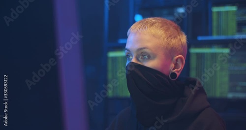 Portrait of Caucasian woman hacker with short hair and masked face sitting in dark room at desk in front of computer. Turning face to camera and taking off mask. Close up of female hacking system. photo