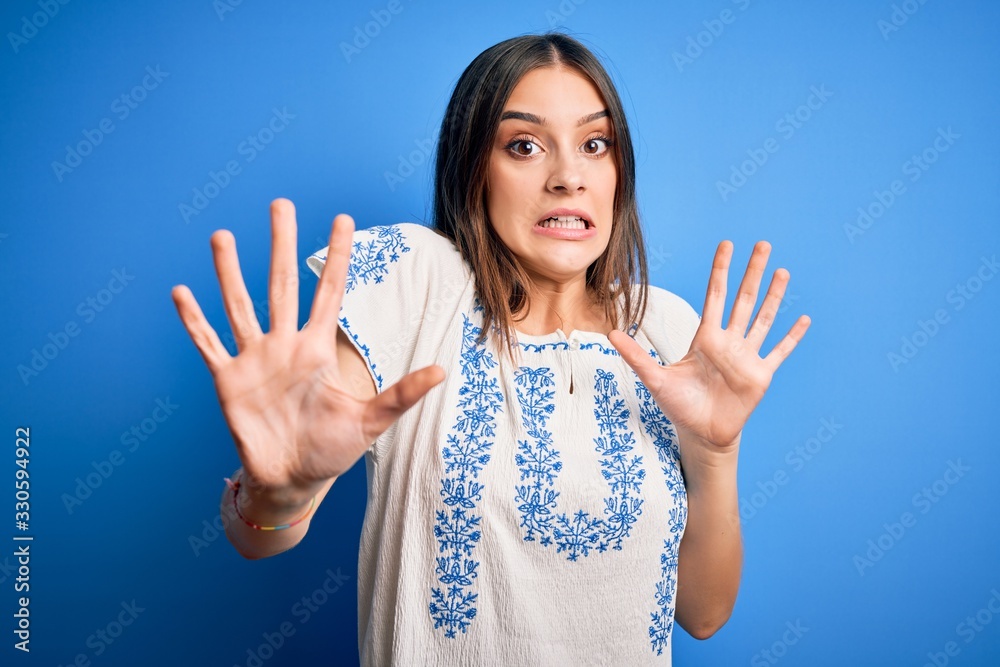 Young beautiful brunette woman wearing casual t-shirt standing over blue background afraid and terrified with fear expression stop gesture with hands, shouting in shock. Panic concept. Young beautiful brunette woman wearing casual t-shirt standing over blue background afraid and terrified with fear expression stop gesture with hands, shouting in shock. Panic concept.
