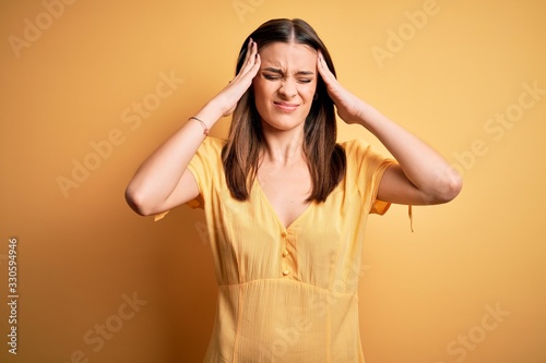 Young beautiful brunette woman wearing casual dress standing over yellow background suffering from headache desperate and stressed because pain and migraine. Hands on head.
