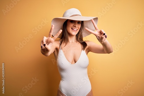 Young beautiful brunette woman on vacation wearing swimsuit and summer hat smiling with tongue out showing fingers of both hands doing victory sign. Number two.
