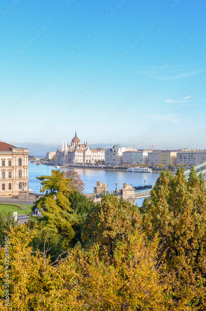Fototapeta premium Amazing cityscape of Budapest, Hungary with Szechenyi Chain Bridge over the Danube River and Hungarian Parliament Building in the background. Trees in the foreground. Vertical photo