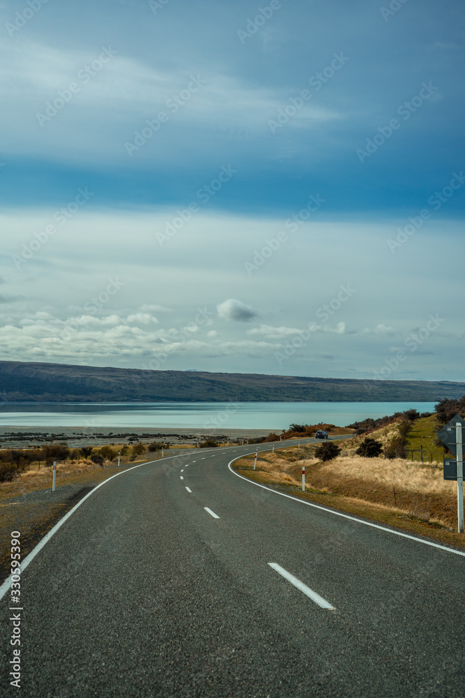 Asphalt road with snowy mountains in the background next to bluish Pukaki Lake, New Zealand