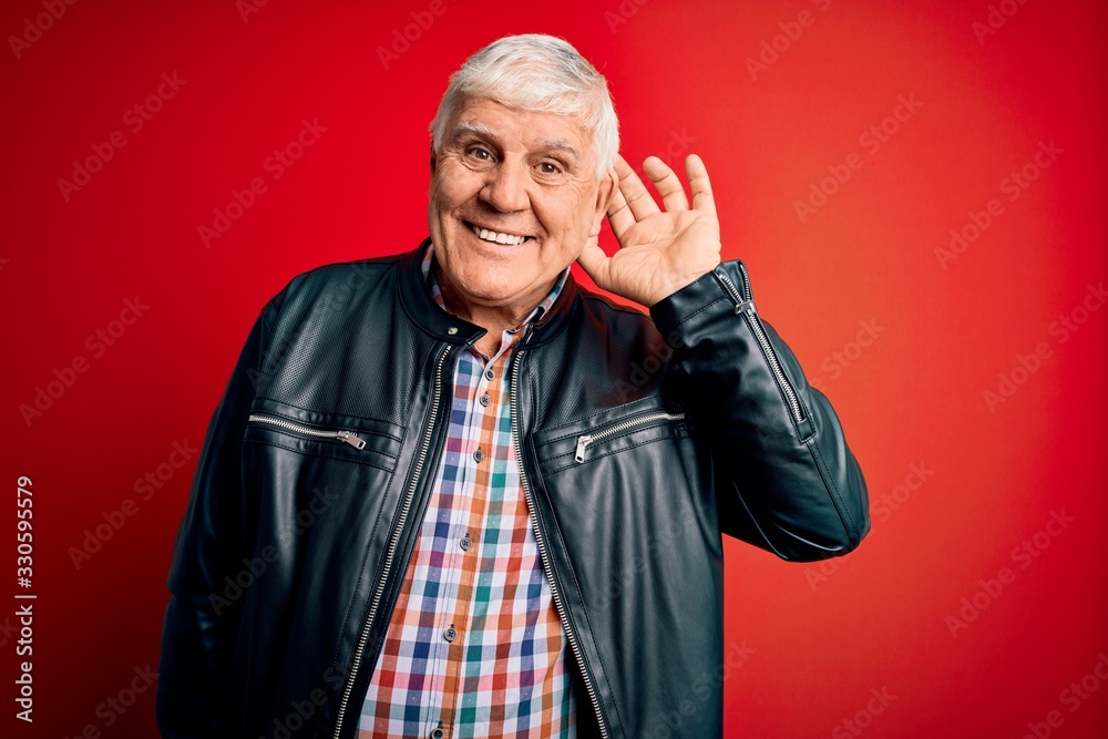 Senior handsome hoary man wearing casual shirt and jacket over isolated red background smiling with hand over ear listening an hearing to rumor or gossip. Deafness concept.
