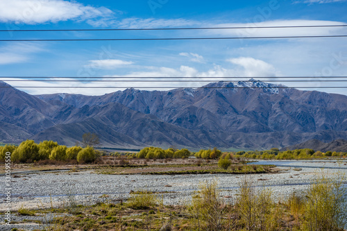 Beautiful dry river with leafy trees on the sides and snow capped mountains in the background taken on a sunny day  New Zealand