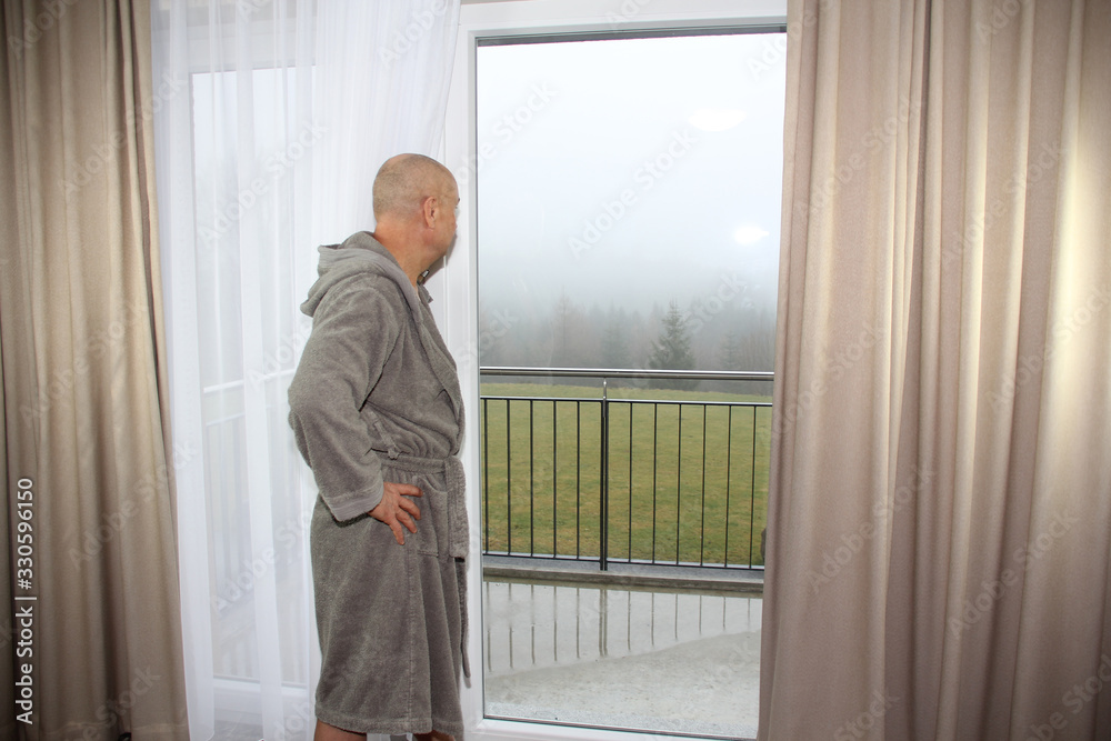Fototapeta premium middle-aged man in a gray bathrobe stands at the window in a hotel room, concept of vacation, travel
