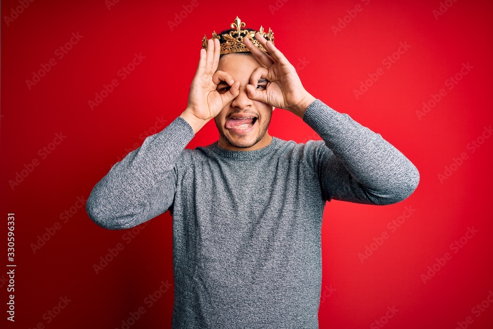 Young handsome man wearing golden crown of prince over isolated red background doing ok gesture like binoculars sticking tongue out, eyes looking through fingers. Crazy expression. Young handsome man wearing golden crown of prince over isolated red background doing ok gesture like binoculars sticking tongue out, eyes looking through fingers. Crazy expression.