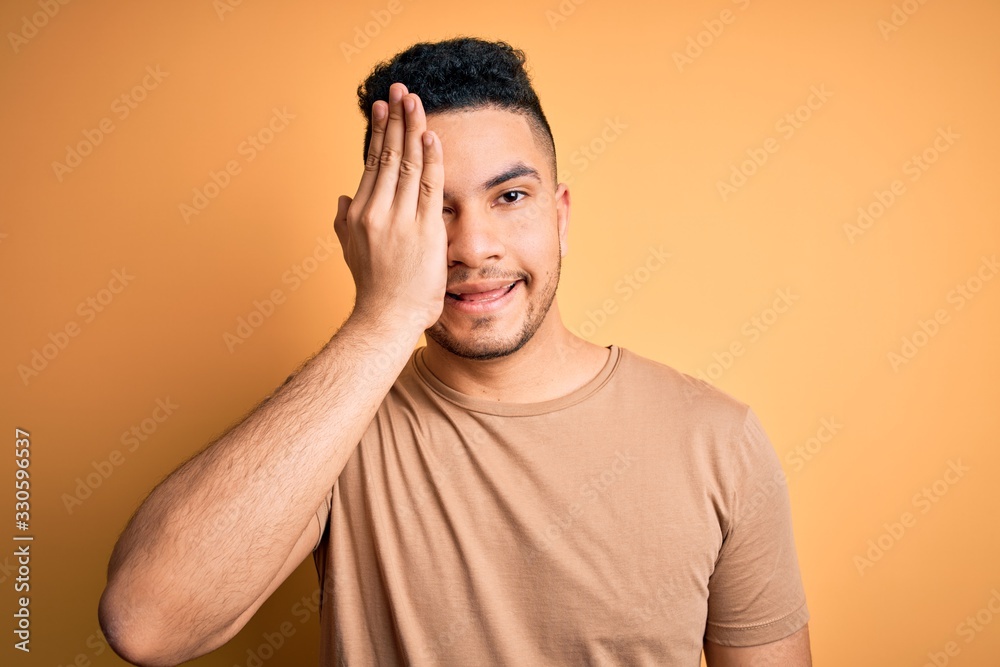 Young handsome man wearing casual t-shirt standing over isolated yellow background covering one eye with hand, confident smile on face and surprise emotion. Young handsome man wearing casual t-shirt standing over isolated yellow background covering one eye with hand, confident smile on face and surprise emotion.