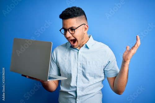 Young handsome businessman wearing glasses working using laptop over blue background very happy and excited, winner expression celebrating victory screaming with big smile and raised hands