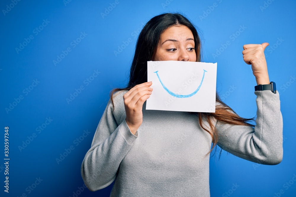 Young beautiful brunette positive woman holding card with smile emotion on mouth pointing and showing with thumb up to the side with happy face smiling Young beautiful brunette positive woman holding card with smile emotion on mouth pointing and showing with thumb up to the side with happy face smiling