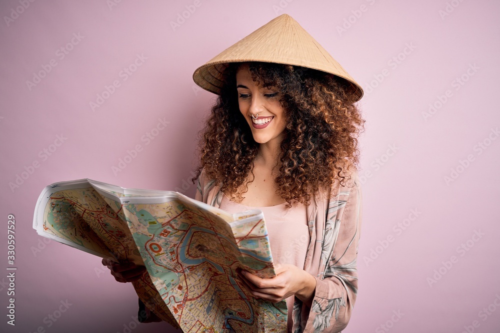 Young beautiful tourist woman with curly hair and piercing wearing asian hat holding city map with a happy face standing and smiling with a confident smile showing teeth