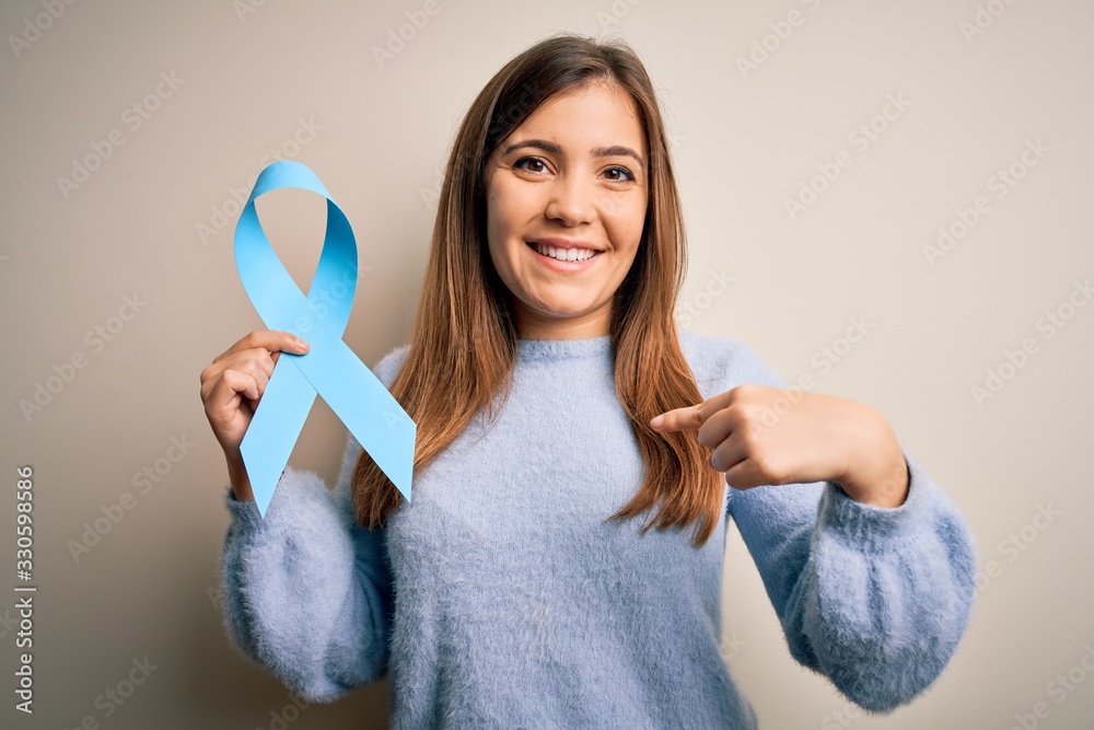 Young blonde woman holding prostate cancer awareness campaing blue ribbon with surprise face pointing finger to himself Young blonde woman holding prostate cancer awareness campaing blue ribbon with surprise face pointing finger to himself