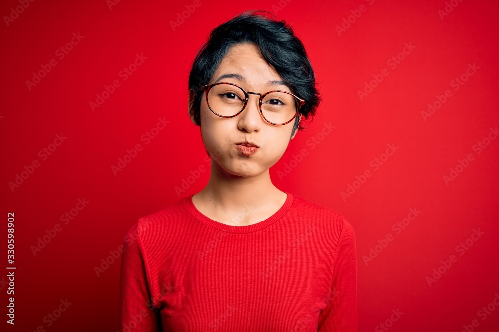 Young beautiful asian girl wearing casual t-shirt and glasses over isolated red background puffing cheeks with funny face. Mouth inflated with air, crazy expression. Young beautiful asian girl wearing casual t-shirt and glasses over isolated red background puffing cheeks with funny face. Mouth inflated with air, crazy expression.