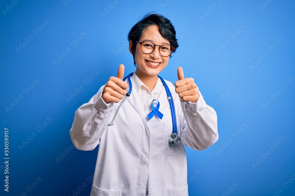 Young beautiful asian doctor girl wearing stethoscope and coat with blue cancer ribbon approving doing positive gesture with hand, thumbs up smiling and happy for success. Winner gesture. Young beautiful asian doctor girl wearing stethoscope and coat with blue cancer ribbon approving doing positive gesture with hand, thumbs up smiling and happy for success. Winner gesture.