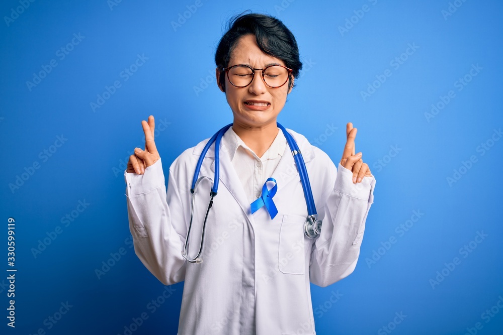Young beautiful asian doctor girl wearing stethoscope and coat with blue cancer ribbon gesturing finger crossed smiling with hope and eyes closed. Luck and superstitious concept. Young beautiful asian doctor girl wearing stethoscope and coat with blue cancer ribbon gesturing finger crossed smiling with hope and eyes closed. Luck and superstitious concept.